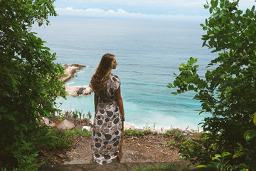 Beautiful and young girl in a dress posing on the shore of the ocean and sea with blue waves. Woman in a long dress model and fashion posing. Relax and relaxation on the island of Bali