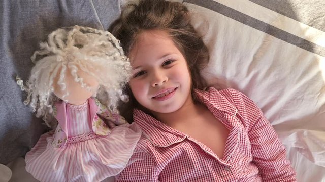 Little girl in pajamas plays in bed with a doll. Fun morning for the child at home in the bedroom. To be at home during the Carntine period. The concept of a happy childhood. 4k video