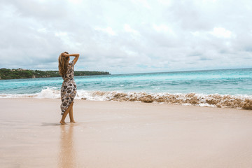 Beautiful and young girl in a dress posing on the shore of the ocean and sea with blue waves. Woman in a long dress model and fashion posing. Relax and relaxation on the island of Bali