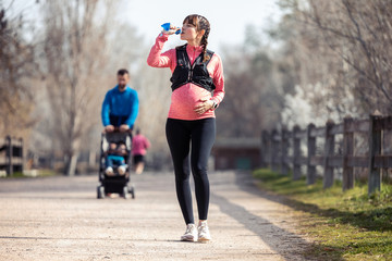 Pregnant young woman drinking water in the park. In the background, her husband walking his son with the cart.