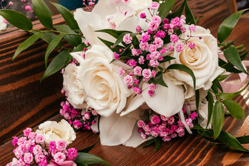 Boutonniere for the groom and wedding bouquet of cream and pink roses, ruskus leaves and pink gypsophila on the brown wooden table.