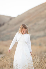 Fototapeta na wymiar A little girl in a white dress outside among the dry grass and foothills
