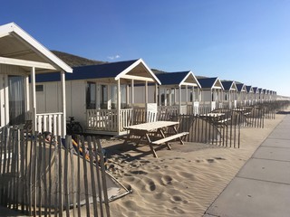 Rental beach houses neatly in row in beach against dunes with table in front and fences, with late afternoon shadows.