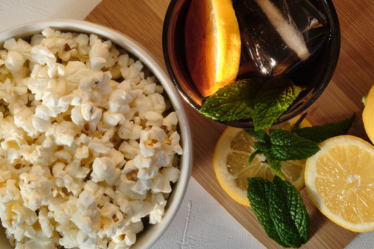 Glass Of Cola With Ice And Lemon And A Bowl Of Popcorn On A White Stone Table. Close Up View From Above. 