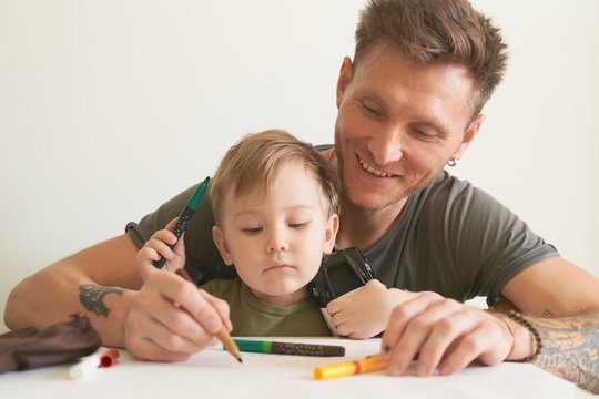 Young father helps son at age of two draw, teaches alphabet. A small boy is sitting on his dads lap near white wall in room at table, on piece of paper writing letters and pictures with felt-tip pen.
