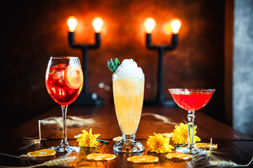 Side view of three different delicious sweet alcohol cocktails in glasses on the wooden table with yellow flowers as decorative elements, horizontal, warm lights