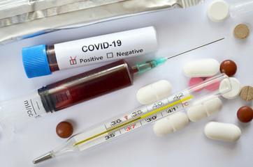 Blood samples in a disposable syringe and test tube with a positive test result for COVID-19, a medical mercury-glass thermometer showing a temperature of 38.2 degrees Celsius and various tablets