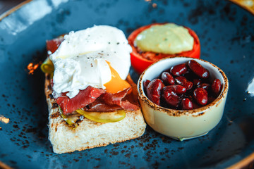 Closeup on appetizing breakfast with squash toasts, beans, poached eggs, meat and vegetables in blue plate on the wooden table, horizontal format