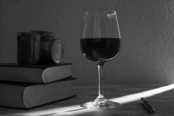 Copa de vino acompañada de unos libros y una camara fotográfica con la luz de la venta entrando por un costado en blanco y negro