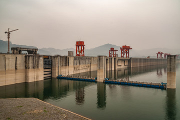 Three Gorges Dam, China - May 6, 2010: Yangtze River. Foggy Morning, Up-river wide long shot along the brown concrete wall with red cranes on top on greenish water. Blue ponton pier.