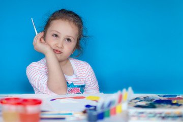 A little girl draws with a brush and colored paints on a white canvas. The cute child looks up thoughtfully. Copy spase. 