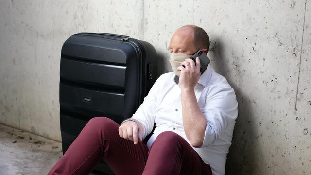 Man waiting his flight at airport terminal, sitting, in waiting room with his trolley bag while is speaking with his smartphone and with mask during Coronavirus pandemic &ndash; Italy 
