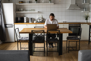 Freelancer working from home on the kitchen and using laptop. Bearded man working with a laptop and reading good news. Handsome successful self entrepreneur sitting and working at his modern home.