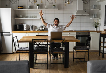 Excited businessman feeling happy about successful project. Man holding hands in the air and looking at laptop while sitting on a kitchen at home. Happy freelancer, student working at laptop at home.
