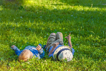 Happy european child shows forefinger up and little brother lying on green grass background. Portrait of child boy and baby boy playing outdoor on lawn. Freedom from ticks and insects. Faceless