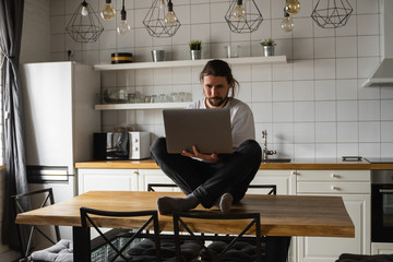 Freelancer working from home sitting on a top of the kitchen table and using laptop. Bearded man working with a laptop and reading news. Successful self entrepreneur working at his modern home.
