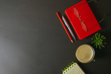 The red notebook stands on a black background, on the notebook there are glasses and next to the notebook, a flower, coffe, pen and a red pencil