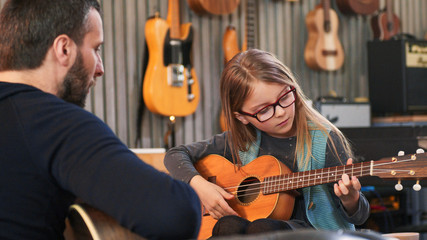 Dad teaching guitar and ukulele to his daughter.Little girl learning guitar at home.Close up.Ukulele class at home. Child learning guitar from her father - obrazy, fototapety, plakaty