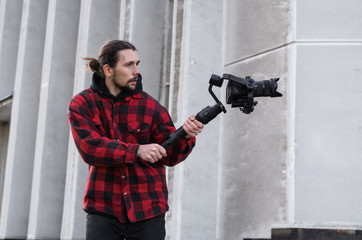 Young Professional videographer holding professional camera on 3-axis gimbal stabilizer. Pro equipment helps to make high quality video without shaking. Cameraman wearing red shirt making a videos.