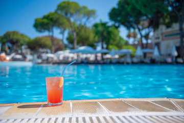 Fototapeta na wymiar Fresh alcoholic red cocktail sex on beach with ice in glass on edge of pool in summer weather against background washed up pool and recreation area. Concept good holiday and travel, place for text.