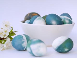 Blue, green, turquoise and white homemade easter eggs colored with a red cabbage natural colour on a white background with blank space decorated with spring flowers in a white bowl closeup.