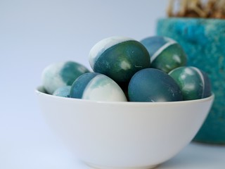 Blue, green, turquoise and white homemade easter eggs colored with a red cabbage natural colour on a white background with blank space decorated with spring flowers in a bowl closeup.
