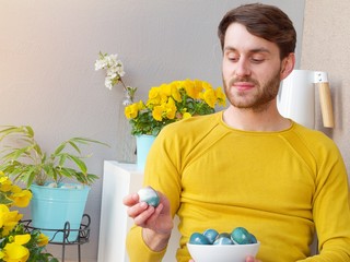 Caucasian man holding a blue and green easter eggs in a bowl during easter time, colored with natural red cabbage coloring, wearing a yellow sweather with yellow flowers in the background.