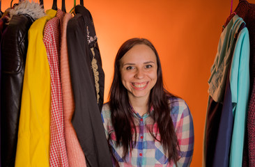 Young woman grimacing standing between clothes in wardrobe. Adult female choosing things in store.