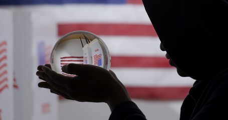 CU silhouetted African American man in a hoodie obscuring his face, looks at crystal ball showing voting booths in front of US flag