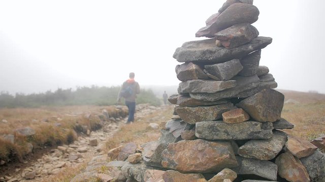 A male mountain hiker walks past a stone cairn trail marker and disappears into thick cloud smoke.