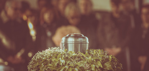 A metal urn with ashes of a dead person on a funeral, with people mourning in the background on a memorial service. Sad grieving moment at the end of a life. Last farewell to a person in an urn.