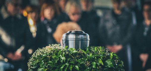 A metal urn with ashes of a dead person on a funeral, with people mourning in the background on a memorial service. Sad grieving moment at the end of a life. Last farewell to a person in an urn. - obrazy, fototapety, plakaty