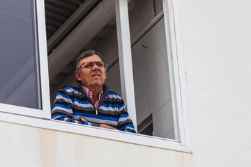 Senior man leaning out the terrace window at home looking at the street. Selective focus.