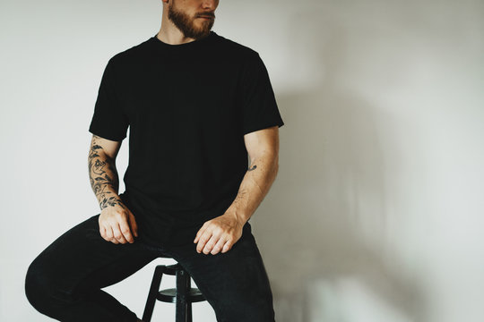 young hipster dressed black t-shirt with empty space for logo, text or design sits on a bar stool against a white wall. mock-up of t-shirt, white wall in the background.