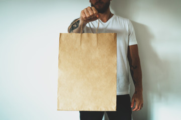 Young hipster man wearing blank white t-shirt and holding craft paper package with empty space for your logo or design, mock-up of shopping bag with handles.