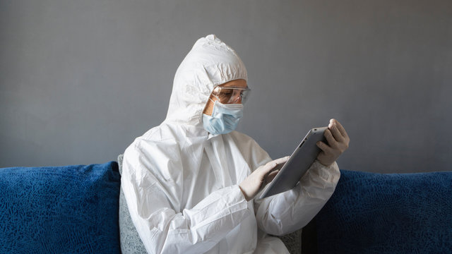 Man in protective suit, medical mask and rubber gloves sits at home and works with tablet on a sofa during quarantine. Designer, artist, architect, businessman at remote work in a pandemic covid.