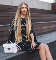 Beautiful young woman with long hair in a black short dress with a small white hand bag. Sits on wooden steps. Summer evening on a city street.