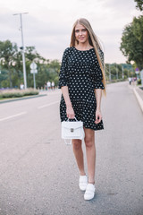 Beautiful young woman with long hair in a black short dress with a small white hand bag. Summer evening en city street.