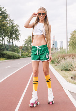 A beautiful long-legged blonde girl in sunglasses, a short white top and green sports shorts rides on roller-bladed roller skates in the summer in a sports park.
