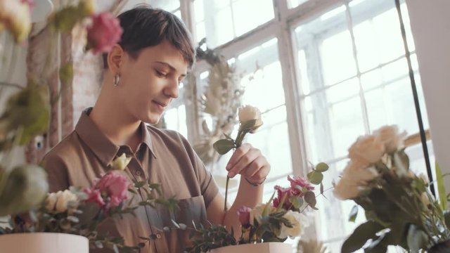 Low angle view of young Caucasian woman cutting rose and putting it into hatbox while working on floral composition in flower shop