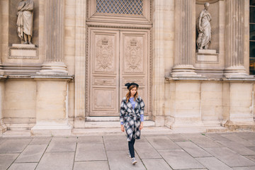 A girl in a beautiful coat stands near the Louvre.