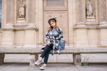Fototapeta na wymiar A girl in a beautiful coat is sitting on a bench near the Louvre.