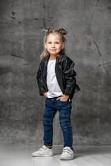 Little smiling blond girl in stylish rock style black leather jacket, blue jeans and white sneakers standing with hands in pockets