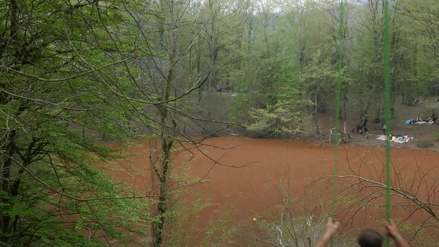 Rear shot of a man swinging at a height in a forest with a red beautiful lagoon in background
