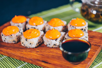 Delicious rolls on wooden board with chopsticks and green tea in teapot on decorative bamboo napkin. Close up of tasty sushi rolls on cutting board on black background.