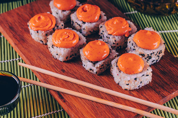 Delicious rolls with spicy sauce on wooden board on decorative bamboo napkin. Close up of tasty sushi rolls with chopsticks on cutting board.