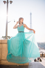 Obraz premium A young beautiful and elegant girl in a blue and green dress stands against the background of the Eiffel tower in Paris.