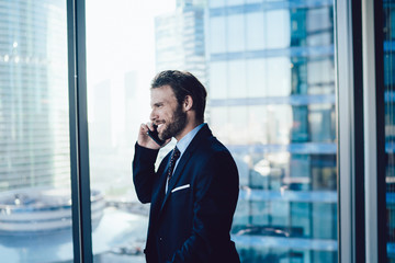 Cheerful bearded male entrepreneur in elegant formal wear having mobile phone conversation about business in office, successful man 30s manager joking talking on cellular near panoramic window