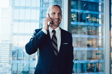 Smiling prosperous businessman in formal outfit standing near office with mobile phone making call for discuss work, confident male entrepreneur dressed in elegant wear enjoying cellular talk