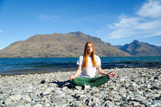 Young Redhead Lady Doing Yoga & Meditating  On A Beach Near A Lake And Mountains